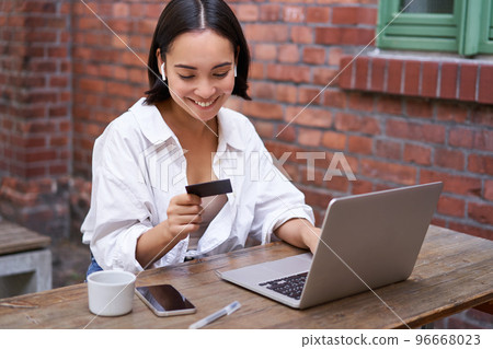 Smiling asian woman with laptop and wireless earphones, paying with credit card, buying online, sitting with cup of coffee Smiling asian woman with laptop and wireless earphones, paying with credit card, buying online, sitting with cup of coffee 96668023