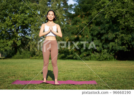 Young mindful woman, meditating on fresh air, doing yoga exercises in park, standing on rubber mat in sportswear Young mindful woman, meditating on fresh air, doing yoga exercises in park, standing on rubber mat in sportswear 96668190