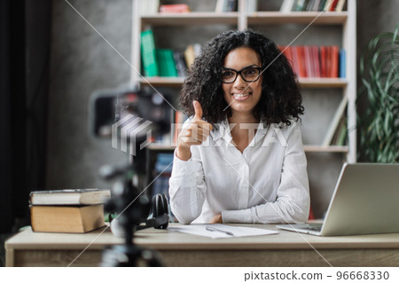 Multiracial woman in white shirt sitting at desk with books, notes and laptop recording video blog Multiracial woman in white shirt sitting at desk with books, notes and laptop recording video blog 96668330