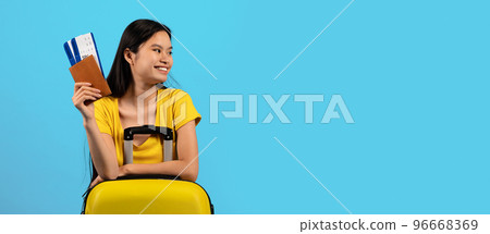 Happy millennial korean lady tourist in yellow t-shirt with suitcase show passport, tickets, looks at empty space 96668369