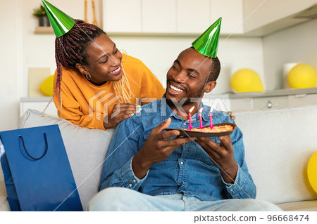 Loving black wife making surprise for her husband, happy man holding birthday pie while sitting on sofa at home Loving black wife making surprise for her husband, happy man holding birthday pie while sitting on sofa at home 96668474