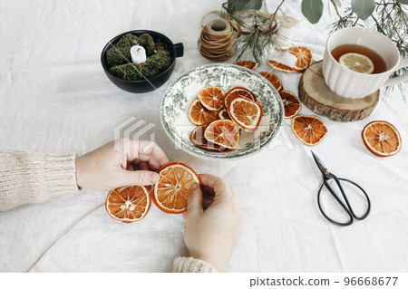 Closeup of womans hands making Christmas garland. Cup of tea, dry oranges fruit and pine, eucalyptus tree branches on white linen table cloth. Festive winter craft table still life. Selective focus. 96668677