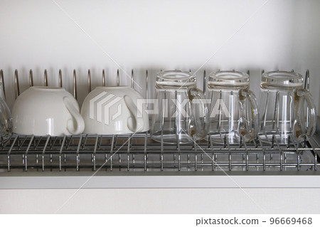 White washed glass and ceramic cups on metal rack inside kitchen cupboard. Modern kitchen with tableware drying White washed glass and ceramic cups on metal rack inside kitchen cupboard. Modern kitchen with tableware drying 96669468