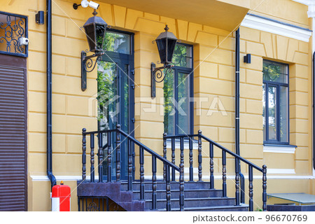 A fragment of a beautiful entrance of the facade of the house with wooden railings on the stairs and openwork lanterns in the old style. A fragment of a beautiful entrance of the facade of the house with wooden railings on the stairs and openwork lanterns in the old style. 96670769