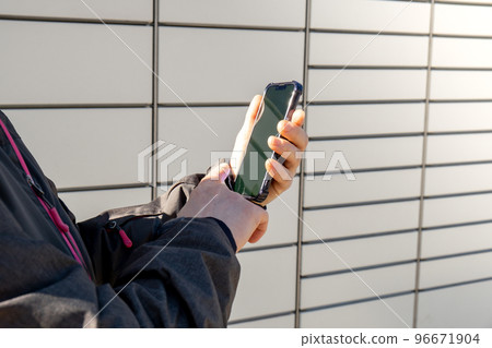 Female hand holding mobile phone next to Parcel locker Collecting parcel from shopping locker. Woman Skans QR Code on Mobile phone Self-service Locker Cell with Bar code Reader Modern Shipping and Female hand holding mobile phone next to Parcel locker Collecting parcel from shopping locker. Woman Skans QR Code on Mobile phone Self-service Locker Cell with Bar code Reader Modern Shipping and 96671904