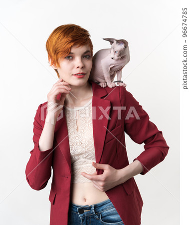 Portrait of hipster redhead young woman with short hair, dressed in red jacket, blue jeans and playful Sphynx Cat sitting on her shoulder. Front view studio shot on white background. Part of series Portrait of hipster redhead young woman with short hair, dressed in red jacket, blue jeans and playful Sphynx Cat sitting on her shoulder. Front view studio shot on white background. Part of series 96674785
