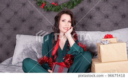 Happy young woman holding gift box for Christmas sitting on bed with xmas decorations, presents. Young girl wearing satin green pajama with holiday decorations at cozy living room. 96675418