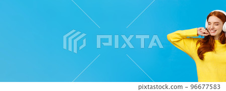 Carefree charming redhead female in yellow sweater, dancing relaxed and joyful with hands lifted up, close eyes and smiling happy, listen music headphones, standing blue background Carefree charming redhead female in yellow sweater, dancing relaxed and joyful with hands lifted up, close eyes and smiling happy, listen music headphones, standing blue background 96677583