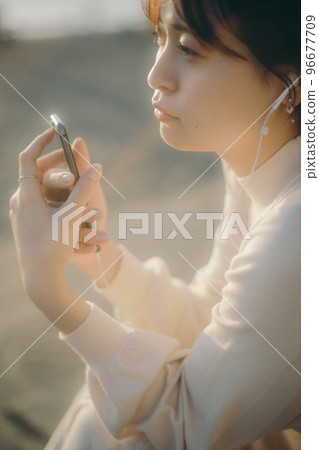 一位年輕女子在智能手機上聽音樂的照片，背景是日落 96677709