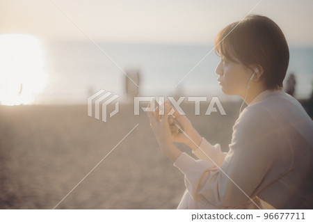 Photo of a young woman listening to music on her smartphone with the sunset in the background Photo of a young woman listening to music on her smartphone with the sunset in the background 96677711