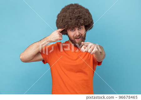 Hey, you are kidding. Man with Afro hairstyle surprised face holding finger near head and pointing finger at camera, stupid gesture. Indoor studio shot isolated on blue background. 96678465