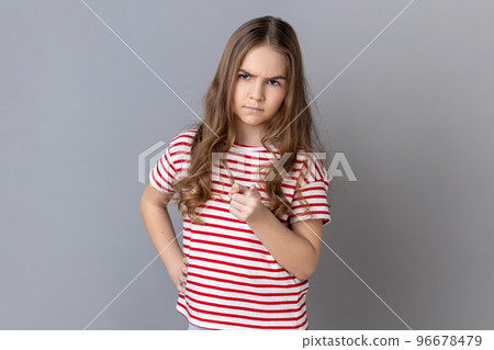 Portrait of little girl wearing striped T-shirt pointing finger at camera and looking with dissatisfied suspicious expression, warning about troubles. Indoor studio shot isolated on gray background. Portrait of little girl wearing striped T-shirt pointing finger at camera and looking with dissatisfied suspicious expression, warning about troubles. Indoor studio shot isolated on gray background. 96678479