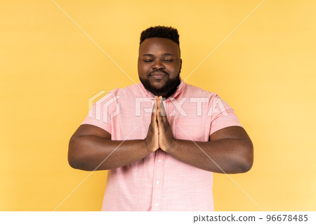 Portrait of man wearing pink shirt keeps hands in yoga gesture, has calm facial expression, keeping palms pressed together and closed eyes. Indoor studio shot isolated on yellow background. 96678485