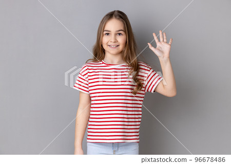 Portrait of adorable cheerful little girl wearing striped T-shirt standing waving hand, looking at camera with engaging toothy smile. Indoor studio shot isolated on gray background. Portrait of adorable cheerful little girl wearing striped T-shirt standing waving hand, looking at camera with engaging toothy smile. Indoor studio shot isolated on gray background. 96678486