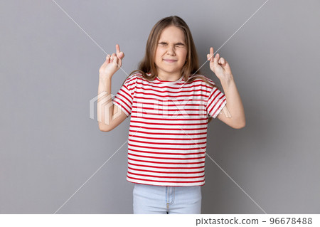Portrait of little girl wearing striped T-shirt holding crossed fingers and wishing for good luck, having hopeful look, hoping for victory. Indoor studio shot isolated on gray background. 96678488