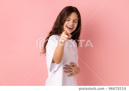 Portrait of positive optimistic joyful little girl wearing white T-shirt laughing out loud holding belly and pointing finger on you, mockery. Indoor studio shot isolated on pink background. 96678519