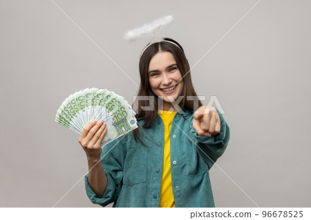 Positive angelic young woman with halo above head holding euro banknotes and pointing to camera, choosing lottery winner, encouraging to earn big money. Indoor studio shot isolated on gray background. Positive angelic young woman with halo above head holding euro banknotes and pointing to camera, choosing lottery winner, encouraging to earn big money. Indoor studio shot isolated on gray background. 96678525
