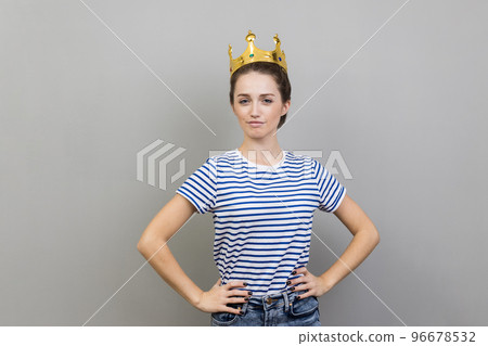 Portrait of woman wearing striped T-shirt and golden crown, looking at camera with confident facial expression, keeps hands on hips. Indoor studio shot isolated on gray background. 96678532