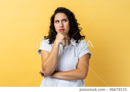 Hmm, I'm need thinking. Portrait of woman with dark wavy hair holding chin and musing, looking away with thoughtful facial expression. Indoor studio shot isolated on yellow background. Hmm, I'm need thinking. Portrait of woman with dark wavy hair holding chin and musing, looking away with thoughtful facial expression. Indoor studio shot isolated on yellow background. 96678535