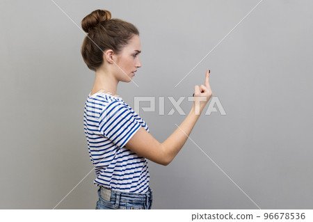 Side view of crazy angry young adult woman wearing striped T-shirt showing middle finger, rude gesture, face full of hatred and resentment. Indoor studio shot isolated on gray background. 96678536
