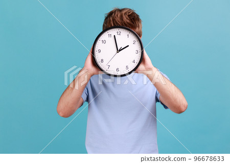 Portrait of unknown anonymous man holding big wall clock hiding his face, time management, schedule and meeting appointment. Indoor studio shot isolated on blue background. 96678633