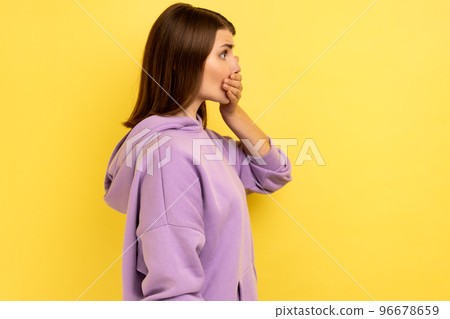 Side view portrait of shocked dark haired woman put hand on mouth and looking ahead, keeping terrible secret, wearing purple hoodie. Indoor studio shot isolated on yellow background. 96678659