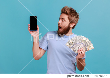 Portrait of surprised amazed handsome bearded man holding dollar banknotes and looking at mobile phone with empty screen, online betting, winning. Indoor studio shot isolated on blue background. 96678666