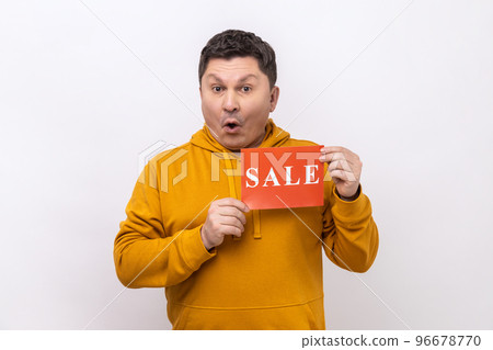 Portrait of astonished man holding sale card in hands looking at camera with open mouth, black Friday, wearing urban style hoodie. Indoor studio shot isolated on white background. Portrait of astonished man holding sale card in hands looking at camera with open mouth, black Friday, wearing urban style hoodie. Indoor studio shot isolated on white background. 96678770