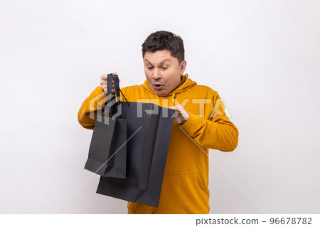 Portrait of middle aged man standing holding black shopping bags and looking inside with shocked facial expression, wearing urban style hoodie. Indoor studio shot isolated on white background. 96678782