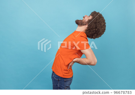 Side view portrait of man with Afro hairstyle wearing orange T-shirt standing with closed eyes, suffering lower back pain, kidney inflammation. Indoor studio shot isolated on blue background. 96678783