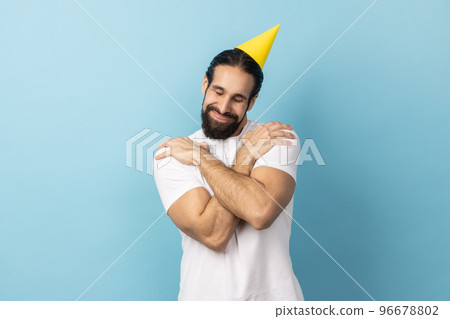 Portrait of smiling handsome birthday man with beard wearing white T-shirt and party cone, celebrating holiday, hugging himself, egoist. Indoor studio shot isolated on blue background. 96678802