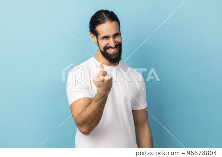Portrait of positive man with beard wearing white T-shirt doing beckoning gesture, calling inviting to approach, smiling and flirting. Indoor studio shot isolated on blue background. Portrait of positive man with beard wearing white T-shirt doing beckoning gesture, calling inviting to approach, smiling and flirting. Indoor studio shot isolated on blue background. 96678803
