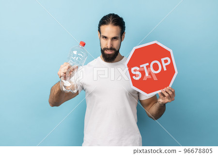 Portrait of strict responsible man with beard wearing white T-shirt holding plastic bottle and stop red sign, worrying about environment. Indoor studio shot isolated on blue background. Portrait of strict responsible man with beard wearing white T-shirt holding plastic bottle and stop red sign, worrying about environment. Indoor studio shot isolated on blue background. 96678805