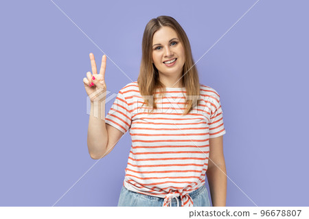 Portrait of beautiful blond woman wearing striped T-shirt smiling happily and showing v sign or peace gesture, rejoicing win. Indoor studio shot isolated on purple background. Portrait of beautiful blond woman wearing striped T-shirt smiling happily and showing v sign or peace gesture, rejoicing win. Indoor studio shot isolated on purple background. 96678807
