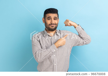 Strong bearded businessman raising hands showing power, pointing at his biceps, feeling independent strong with proud look, wearing striped shirt. Indoor studio shot isolated on blue background. Strong bearded businessman raising hands showing power, pointing at his biceps, feeling independent strong with proud look, wearing striped shirt. Indoor studio shot isolated on blue background. 96678808