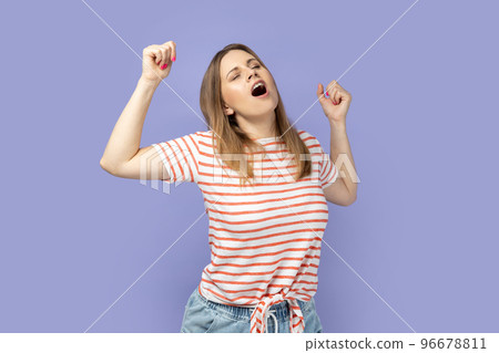 Portrait of tired blond woman wearing striped T-shirt standing yawning with closed eyes, stretching arms, feeling exhausted need rest. Indoor studio shot isolated on purple background. 96678811