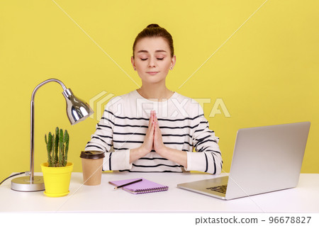 Open minded woman office worker in striped shirt pressing hands together meditating at workplace, preparing herself for work. Indoor studio studio shot isolated on yellow background. Open minded woman office worker in striped shirt pressing hands together meditating at workplace, preparing herself for work. Indoor studio studio shot isolated on yellow background. 96678827