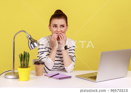 Anxious young woman office manager biting her nails sitting at workplace and looking at camera with nervous worry expression. Indoor studio studio shot isolated on yellow background. Anxious young woman office manager biting her nails sitting at workplace and looking at camera with nervous worry expression. Indoor studio studio shot isolated on yellow background. 96678828