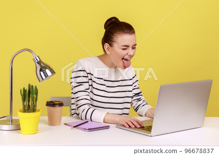 Playful funny woman office worker in striped shirt showing tongue out, expressing positive childish emotions, chatting via web camera on laptop. Indoor studio studio shot isolated on yellow background 96678837