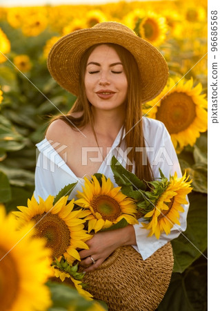 A girl in a hat on a beautiful field of sunflowers against the sky in the evening light of a summer sunset. Sunbeams through the flower field. Natural background. A girl in a hat on a beautiful field of sunflowers against the sky in the evening light of a summer sunset. Sunbeams through the flower field. Natural background. 96686568