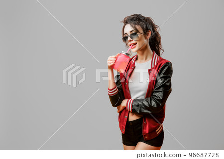 Cute curly stylish young woman in sunglasses drinking beverage in glass over gray background 96687728