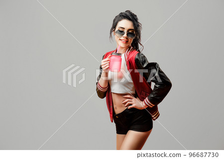 Cute curly stylish brunette girl in sunglasses holds beverage in glass over gray background 96687730