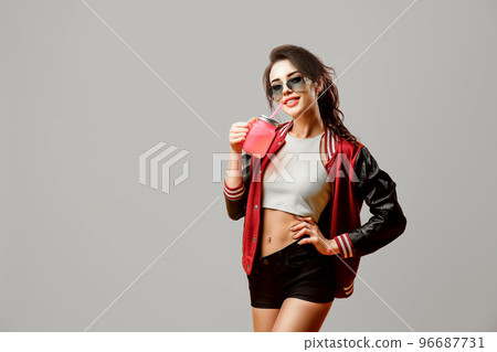 Smiling curly stylish brunette girl in sunglasses holds beverage in glass over gray background 96687731