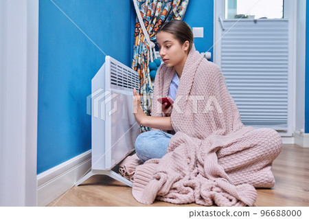 Young female sitting near an electric heater at home 96688000