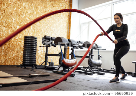 Attractive young slim and fit sportswoman working out in the gym for functional training doing crossfit exercises with battle ropes Attractive young slim and fit sportswoman working out in the gym for functional training doing crossfit exercises with battle ropes 96690939