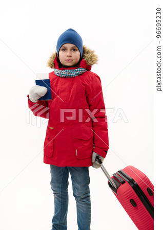 Full length portrait of teenage traveler boy wearing down jacket and blue jeans, going for winter vacations, walking with boarding pass and red suitcase over white background. Journey. Travel concept 96695230