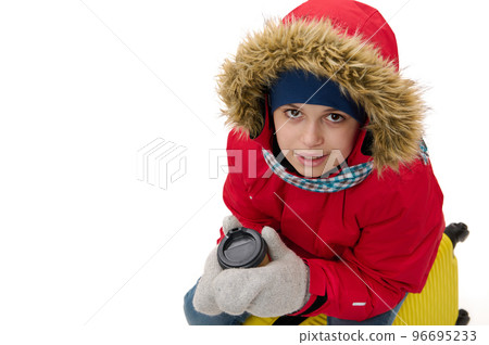 Charming teenager traveler boy in red parka with fur hood and wool mittens, warms his hands with hot drink in disposable paper cup, looks at camera, sitting on suitcase, isolated on white background Charming teenager traveler boy in red parka with fur hood and wool mittens, warms his hands with hot drink in disposable paper cup, looks at camera, sitting on suitcase, isolated on white background 96695233
