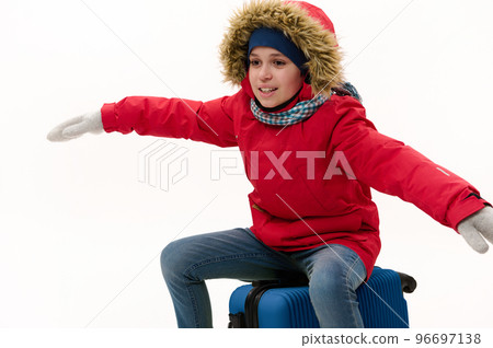 Teenage boy in red parka with fur hood, sits on blue suitcase, stretches his arms to the sides, imitating airplane during flight, isolated on white background. Winter travel concept. Holiday. Vacation Teenage boy in red parka with fur hood, sits on blue suitcase, stretches his arms to the sides, imitating airplane during flight, isolated on white background. Winter travel concept. Holiday. Vacation 96697138