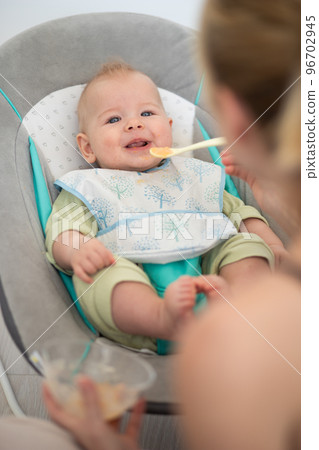 Mother spoon feeding her baby boy child in baby chair with fruit puree. Baby solid food introduction concept Mother spoon feeding her baby boy child in baby chair with fruit puree. Baby solid food introduction concept 96702945