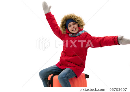 Caucasian smiling teenage traveler boy, wearing warm red down jacket, wool mittens and blue jeans, sitting on orange suitcase, travels abroad, isolated over white. Kids travelling on weekends getaway Caucasian smiling teenage traveler boy, wearing warm red down jacket, wool mittens and blue jeans, sitting on orange suitcase, travels abroad, isolated over white. Kids travelling on weekends getaway 96703607